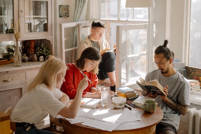 Four student roommates sit in a brightly lit kitchen and study around a kitchen table littered with papers and books