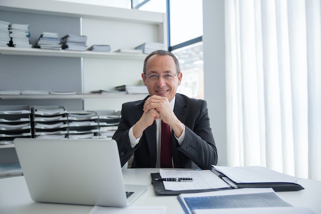Smiling person in a suit and glasses sitting behind a desk